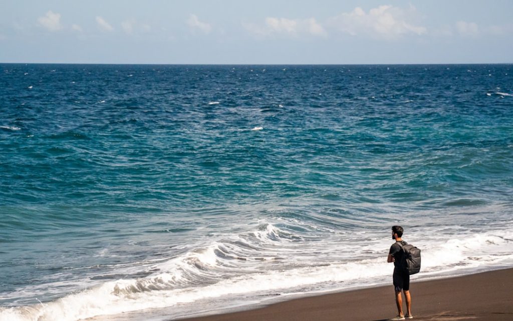 Man with backpack standing on dark sand beach. traveler using smartphone on beach tropical destination