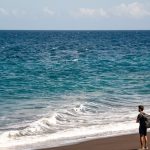 Man with backpack standing on dark sand beach. traveler using smartphone on beach tropical destination