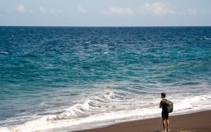 Man with backpack standing on dark sand beach. traveler using smartphone on beach tropical destination