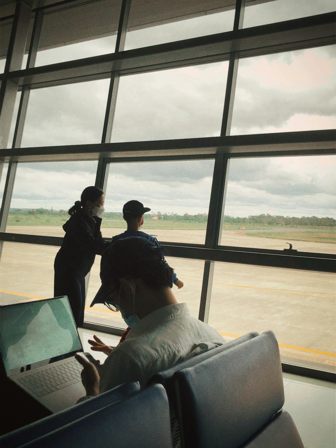 a couple of people standing in front of a window person using laptop in airport, streaming video, travel suitcase nearby