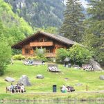 a group of people sitting on a bench in front of a lake traditional gathering, cultural clothing, mountain landscape, community celebration