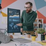 A man pointing at a vertical monitor displaying data charts to two colleagues creative review dashboard, marketing team collaboration, online proofing interface