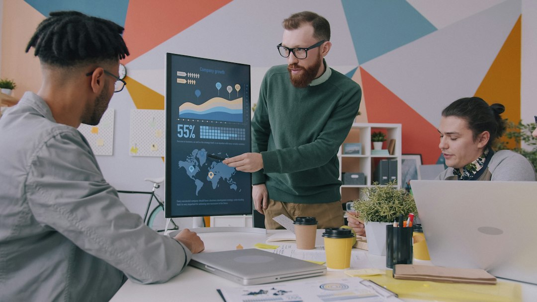 A man pointing at a vertical monitor displaying data charts to two colleagues creative review dashboard, marketing team collaboration, online proofing interface