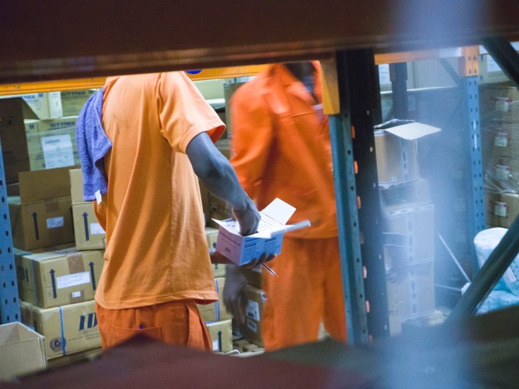 standing man in orange t-shirt holding white box warehouse shelves inventory boxes workers logistics center