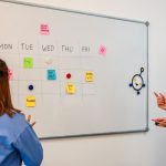 two women standing in front of a whiteboard with sticky notes on it creative review dashboard, marketing team collaboration, online proofing interface