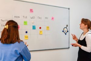 two women standing in front of a whiteboard with sticky notes on it creative review dashboard, marketing team collaboration, online proofing interface