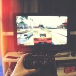 person holding Sony PS3 controller in front of flat screen monitor handheld console gaming screen, high resolution gameplay, vivid graphics display