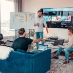 a group of people sitting around a living room project management dashboard analytics screen, team video call on monitor, digital collaboration tools, remote software team