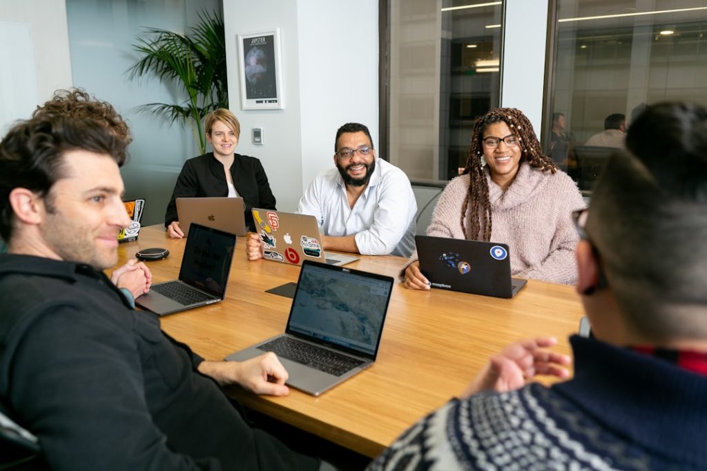 four people all on laptops, two men and two women, listen to person talking in a board meeting  digital collaboration workspace laptops strategy meeting