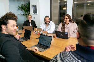 four people all on laptops, two men and two women, listen to person talking in a board meeting  digital collaboration workspace laptops strategy meeting