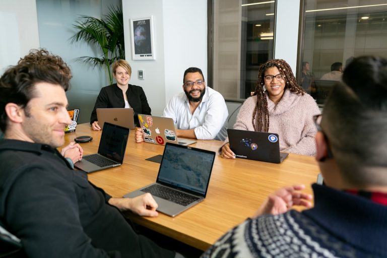 four people all on laptops, two men and two women, listen to person talking in a board meeting  digital collaboration workspace laptops strategy meeting