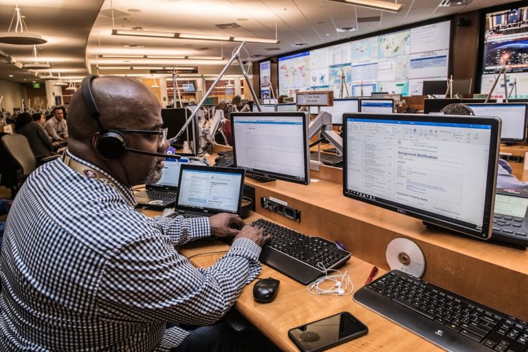 man in black and white checkered dress shirt using computer emergency operations center, multiple monitors, crisis response team