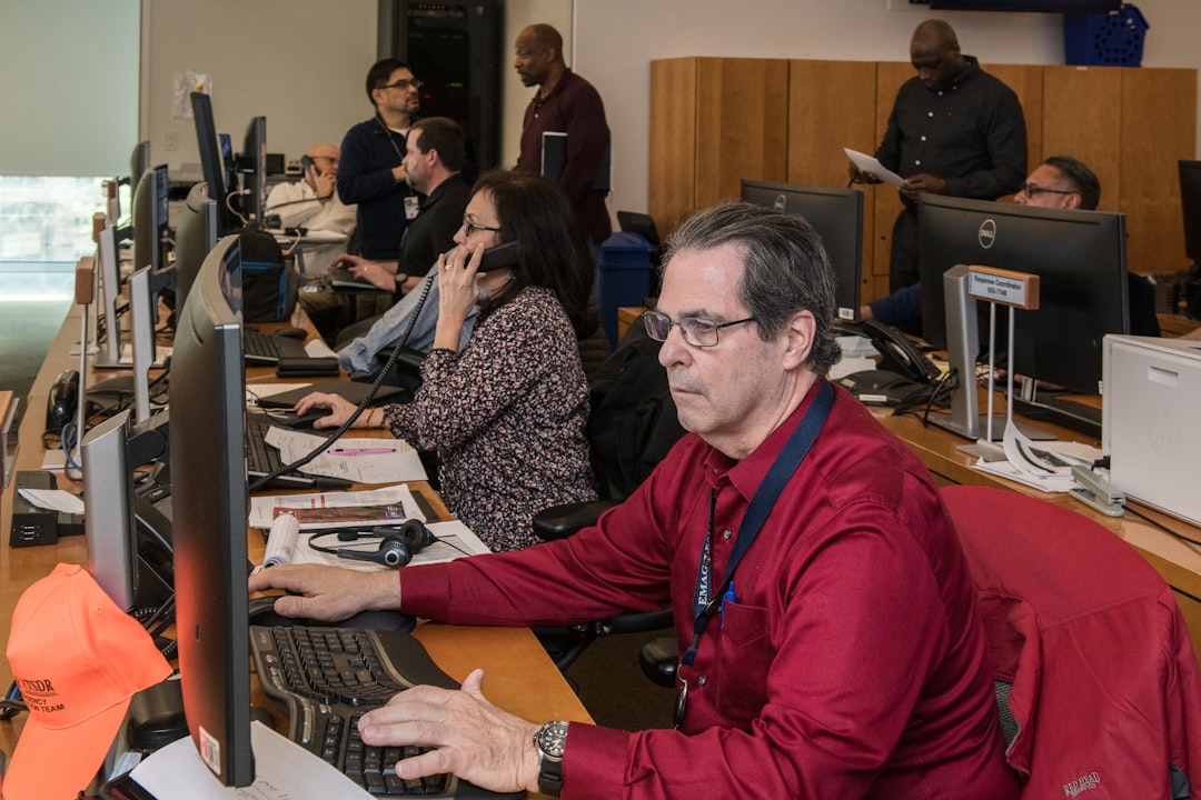 man in red jacket sitting beside woman in black and white long sleeve shirt emergency operations center, multiple monitors, crisis response team