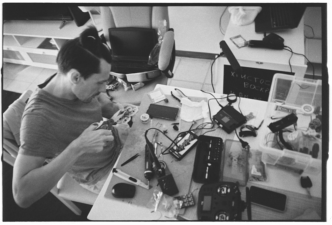 Man working on electronics at a cluttered desk drone battery charging station, battery health screen, drone maintenance workspace