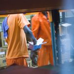 standing man in orange t-shirt holding white box warehouse shelves inventory boxes workers logistics center