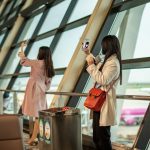 Two women taking selfies by airport window traveler swapping sim card, smartphone close up, airport setting