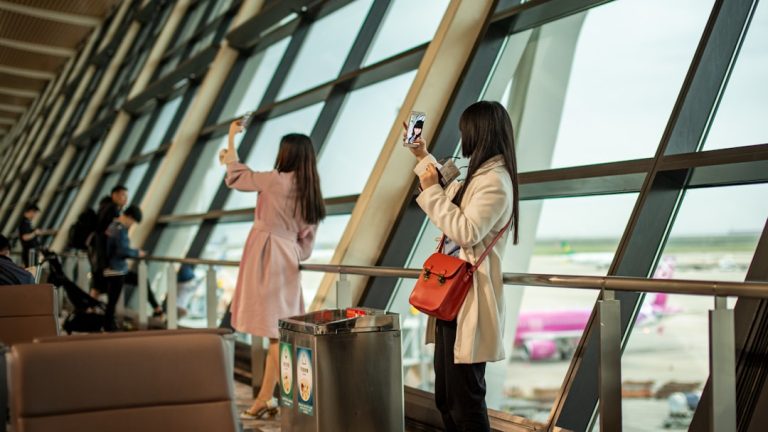 Two women taking selfies by airport window traveler swapping sim card, smartphone close up, airport setting