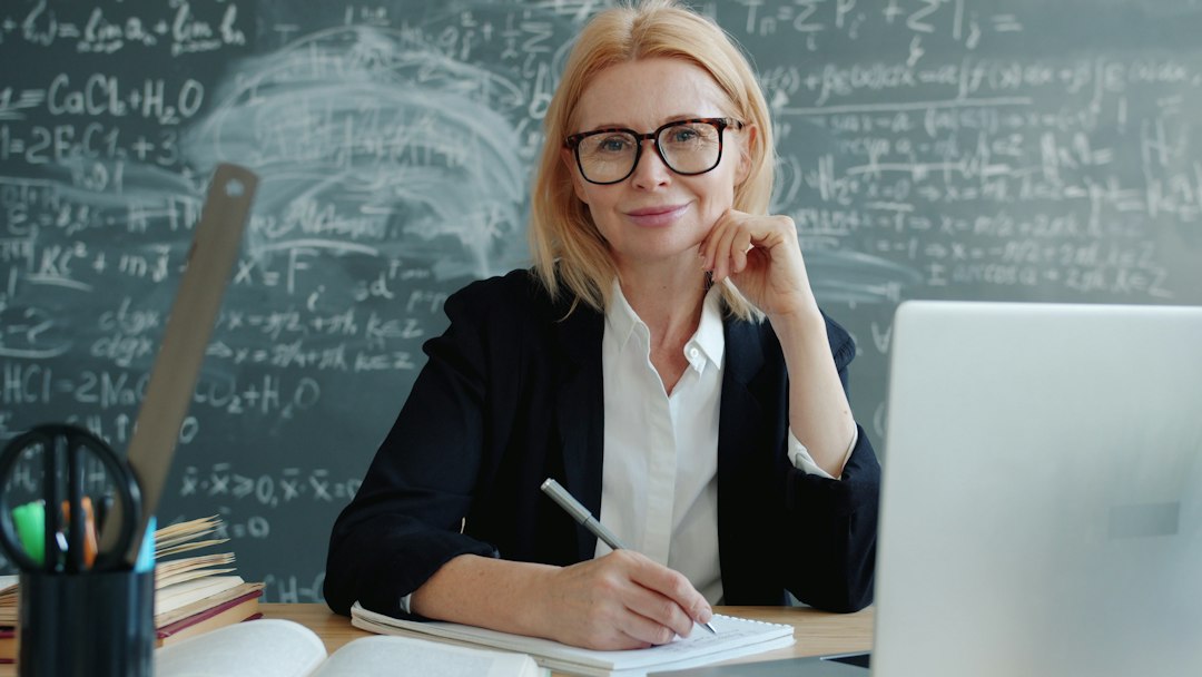 A female teacher smiling at the desk with a laptop teacher laptop, gradebook, classroom