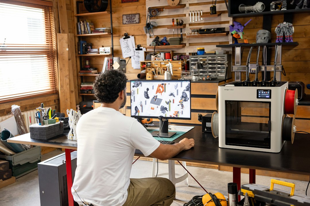 Man working on computer in a workshop with 3D printer cad comparison, drafting software, design tools